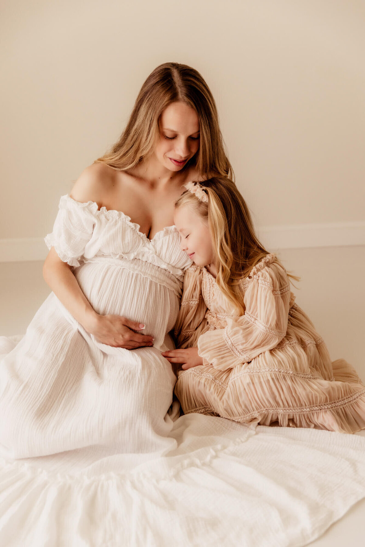 Girl smiles while snuggling her pregnant mother while sitting on white flooring.