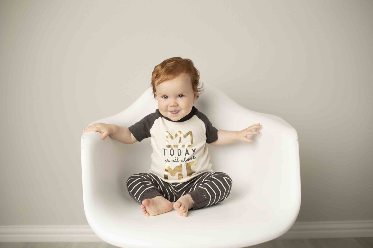 Redhead birthday boy smiling as he sits in a white chair in the studio, surrounded by soft lighting