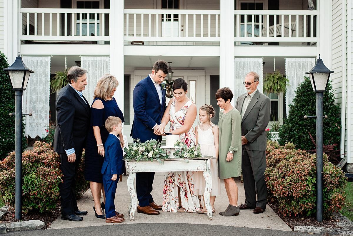 The bride and groom stand at a small white table with their family on either side of them as they cut their wedding cake in front of a large white mansion with a double porch. The table is decorated in white flowers and greenery. The small one tier cake is on a white pedestal stand with a white flower and greenery decorating it. To the left the groom's parents are wearing a black suit and dark blue dress. To the right the bride's parents wear a gray suit and light green dress. The  flower girl is wearing a white sundress and the groom and ring bearer have matching cobalt suits. The bride is wearing a sleeveless white gown with a pink floral pattern. The bride and groom hold hands as they cut into their wedding cake.