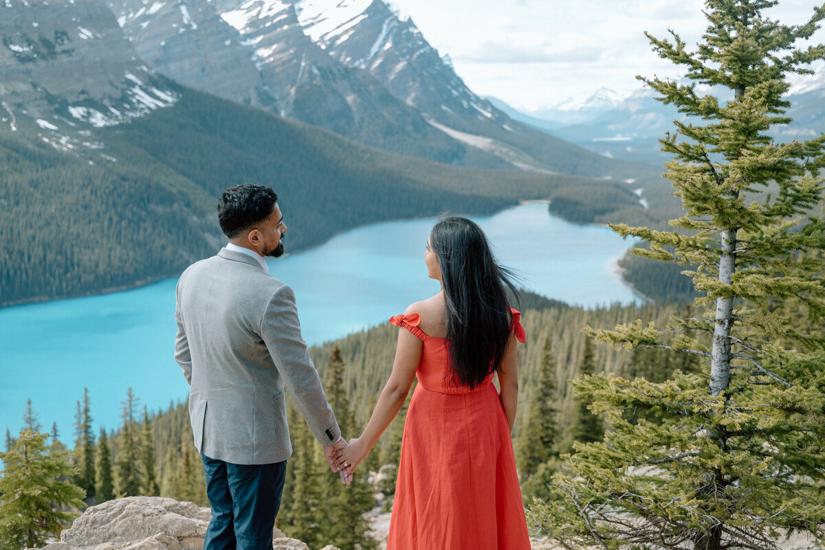 Couple walking hand-in-hand along Moraine Lake shoreline with snowy peaks behind.