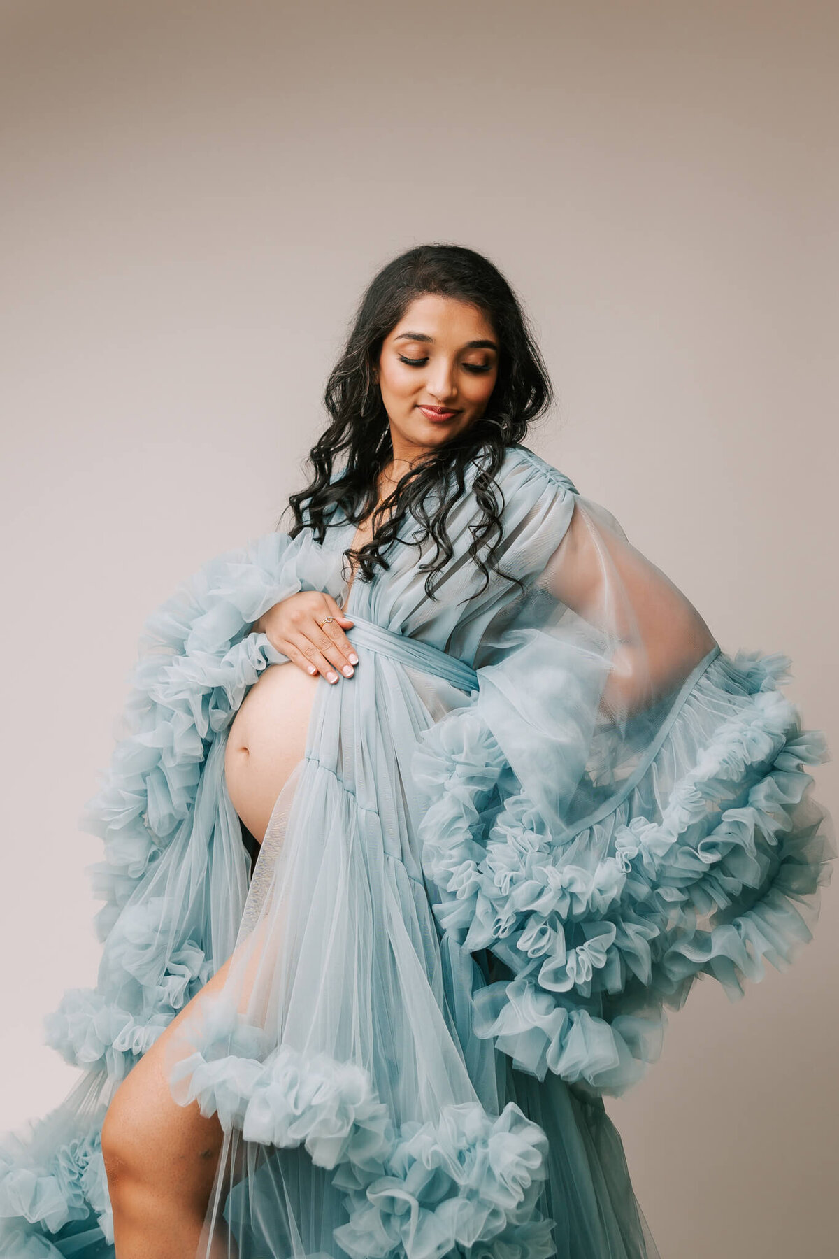 beautiful portrait of a woman in studio with her hand on her pregnant belly. She has black hair and is looking to the right over her shoulder.