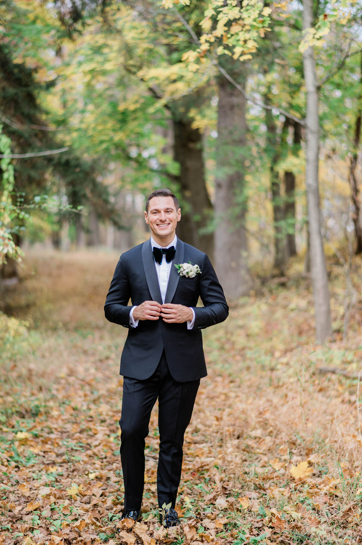 The groom stands confidently in a wooded area, smiling at the camera in his classic black tuxedo with a white floral boutonnière