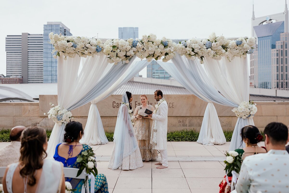 White and blue mandap with white floral decor sits in front of the Nashville skyline. The bride is wearing a white beaded saree with a light blue dupatta and the groom is wearing a white sherwani. The officiant, dressed in light gold, stands by as they exchange their vows at their Nashville Indian Wedding.