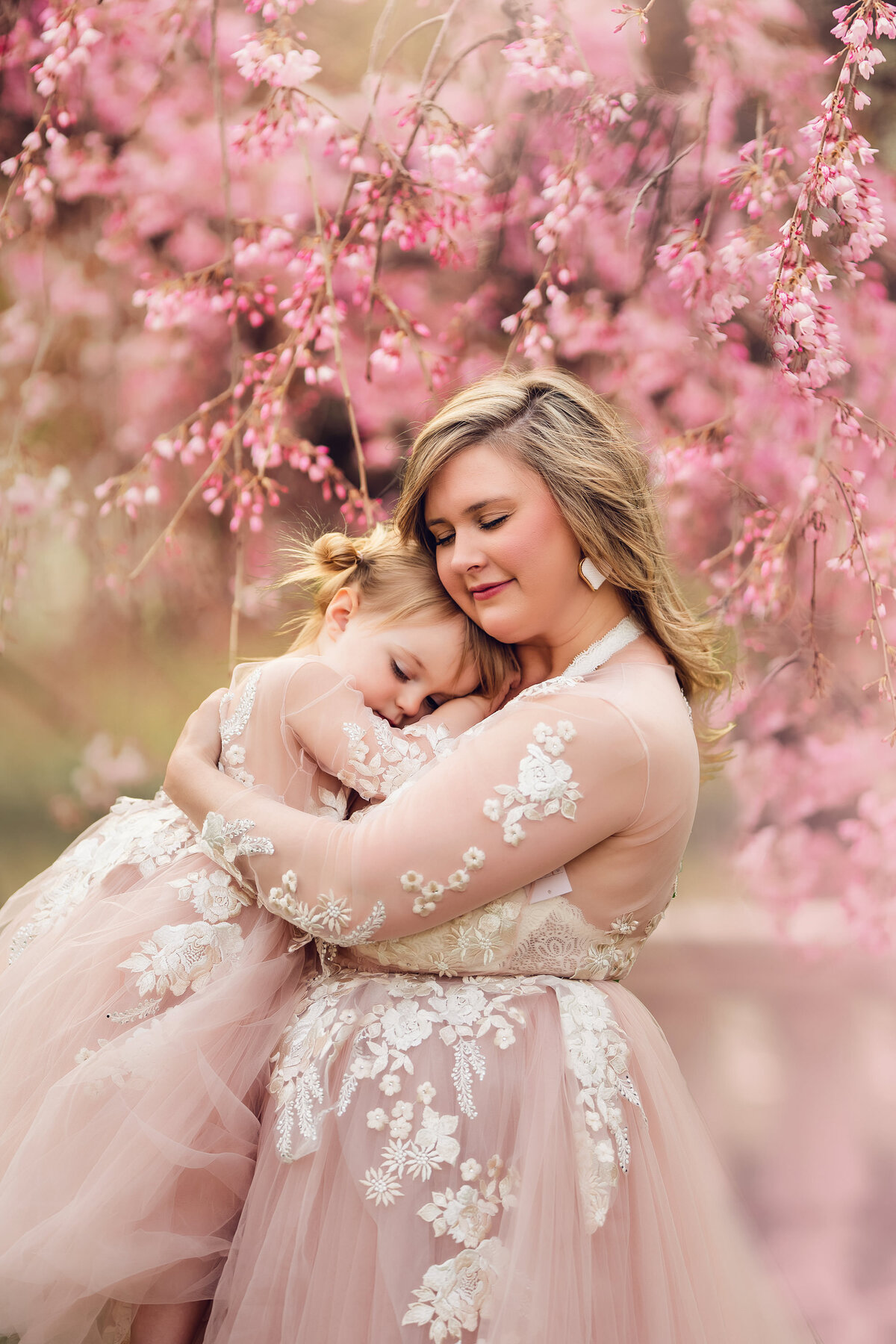 A mother and her young daughter wearing matching, blush gowns embrace surrounded by trees covered in pink blossoms.