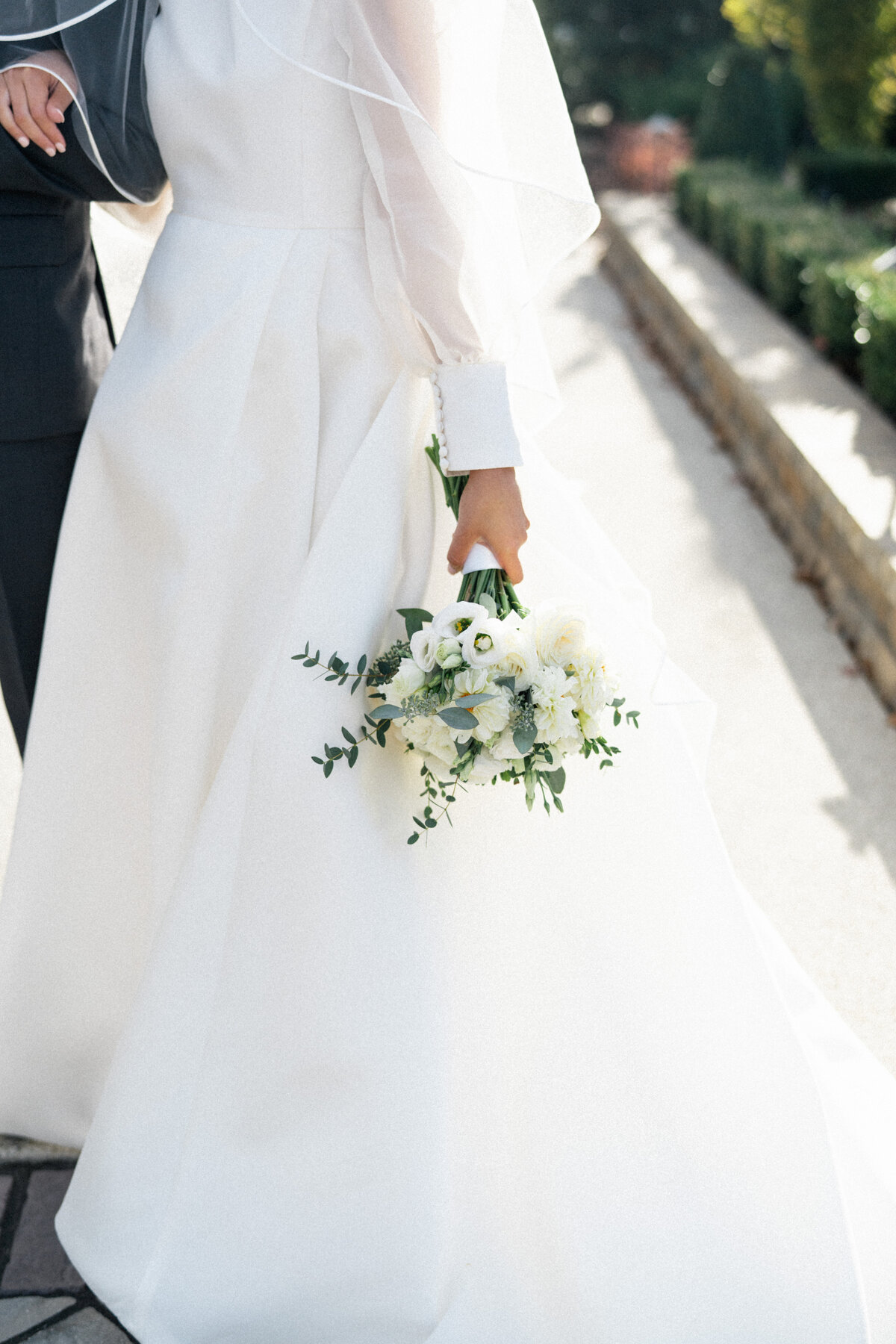 Close-up of a bride in an elegant white gown holding a bouquet of white and green flowers, walking outdoors on a sunny day.