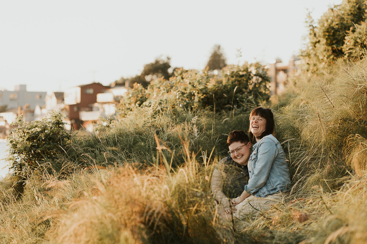 A couple sits in tall grass, leaning against each other and laughing.