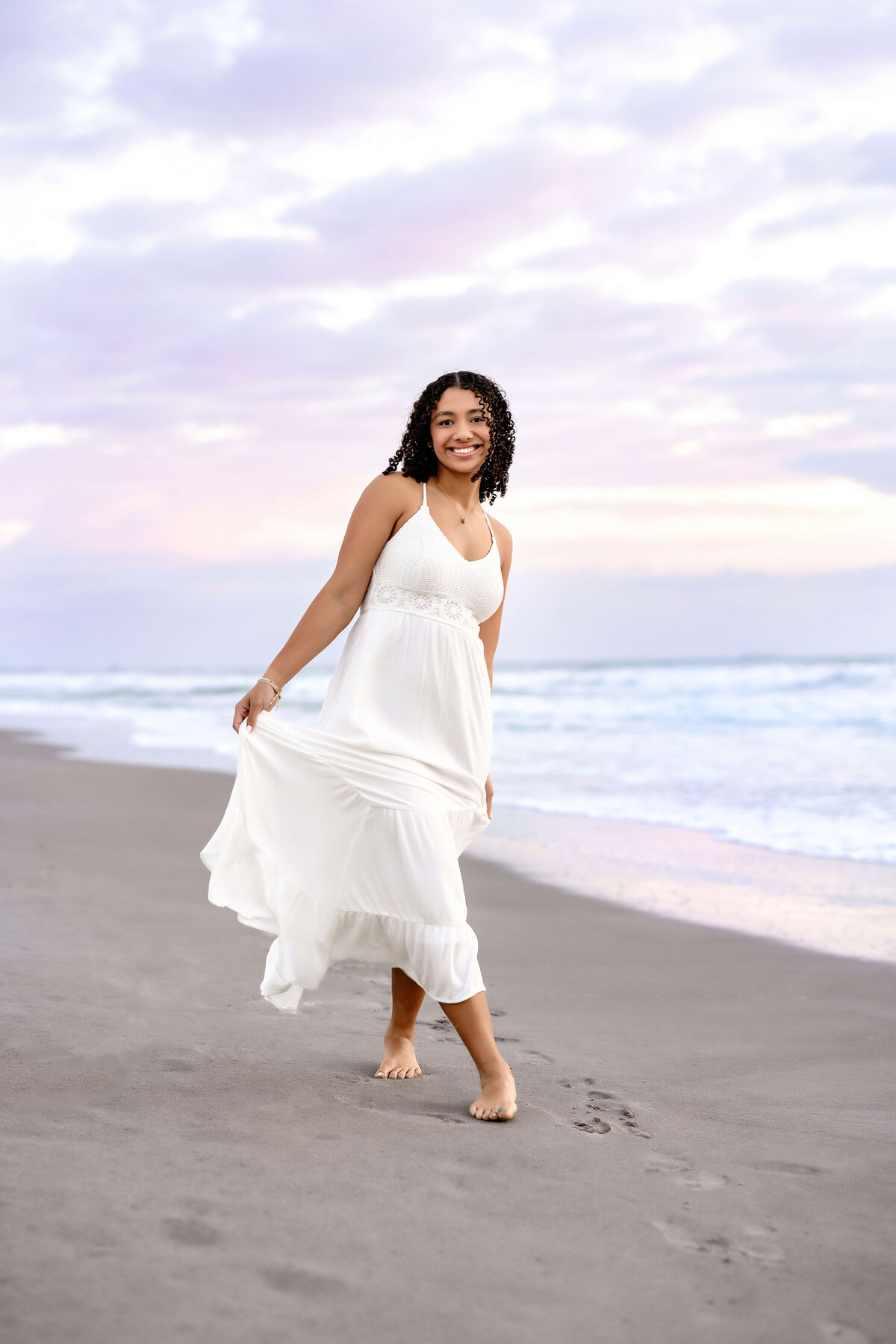 Orlando senior photographer capturing a teen girl in a flowy white dress, walking toward the camera and smiling against a soft pastel cotton candy sky.