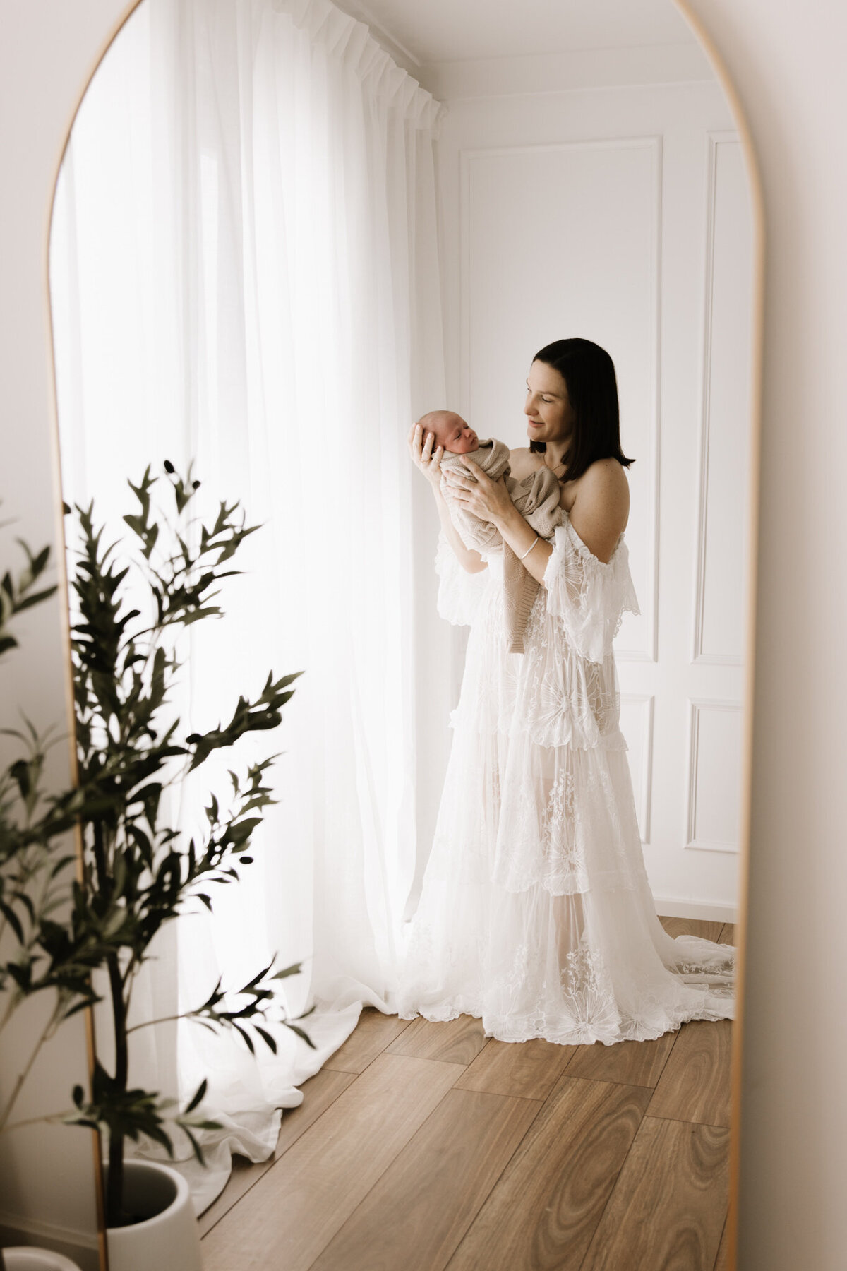 Mother holding her newborn baby near sheer curtains in a soft, light-filled Southwest Sydney photography studio, captured through a mirror with elegant styling.