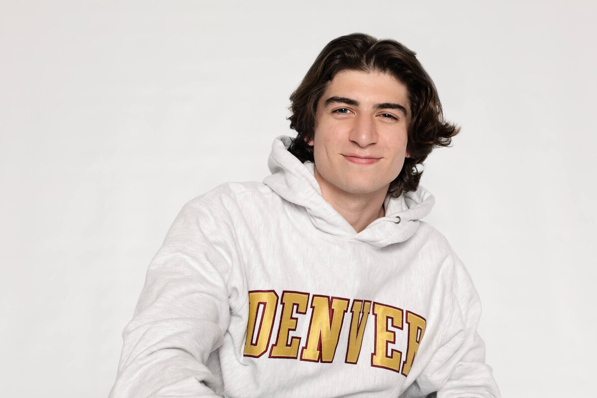 A person with brown hair is smiling while wearing a white hoodie with DENVER written in yellow and red letters. The background is plain and light-colored.