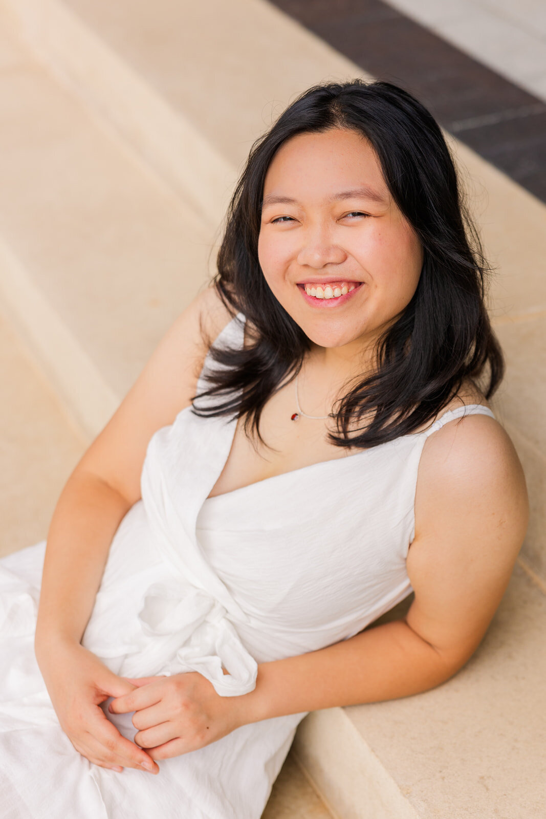 teenage girl wearing a white short dress sitting on stairs resting back on her elbow smiling at the camera during senior photoshoot with a neutral background