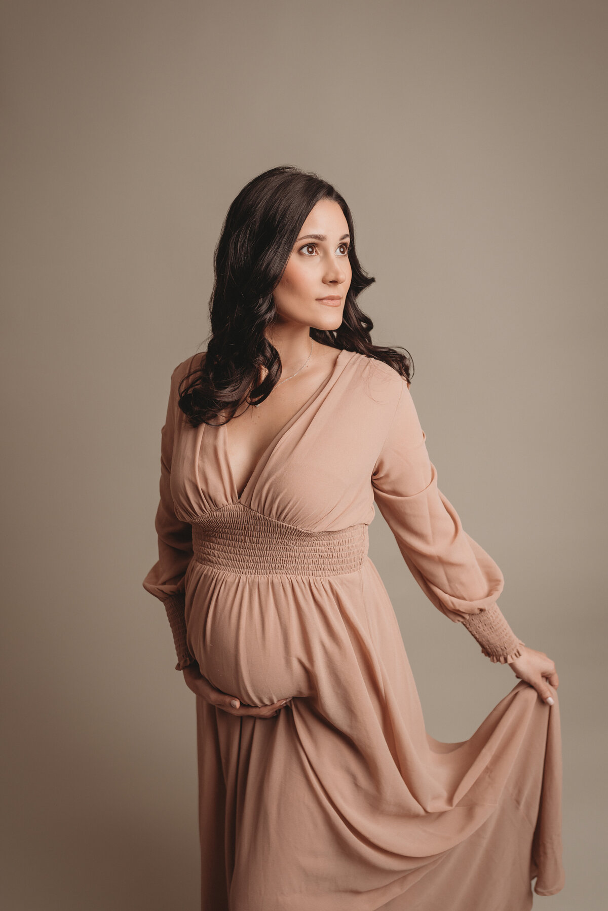 Pregnant woman wearing flowing light pink long sleeve V neck dress holding baby bump with one hand and dress with other hand looking up and to the side