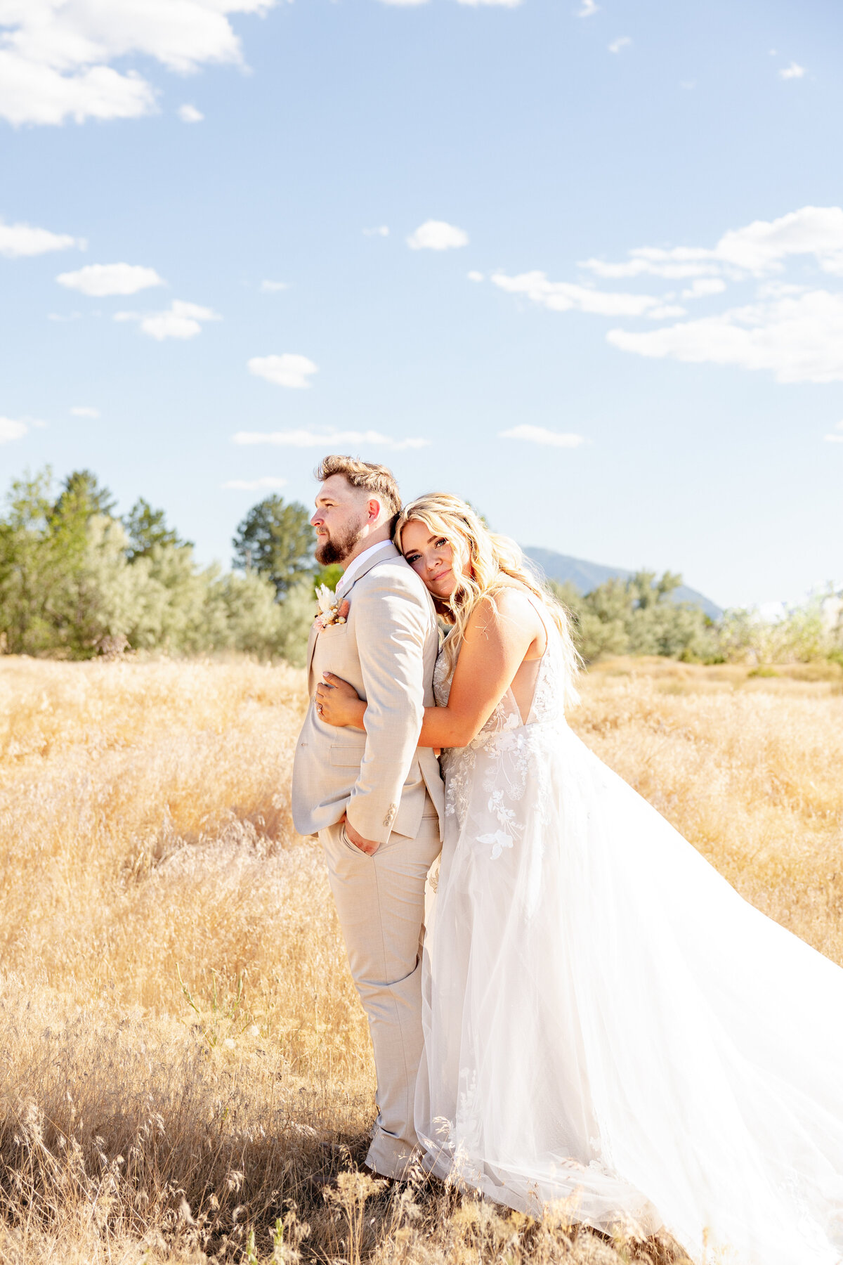 The bride gently rests her head on the groom’s back in an open field, as golden grass and a mountain view create a peaceful backdrop.