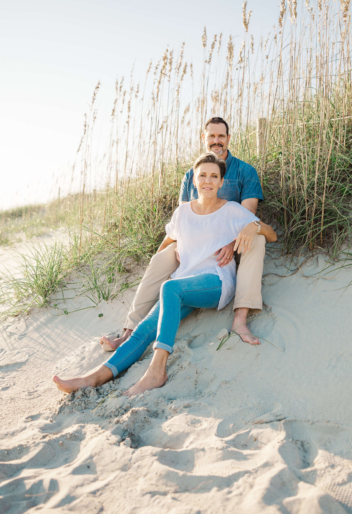 A couple sits on a sandy beach near tall grasses, captured by a talented family photographer in Fayetteville, NC. The woman is seated in front of the man, both barefoot and dressed casually, savoring the sunny day.