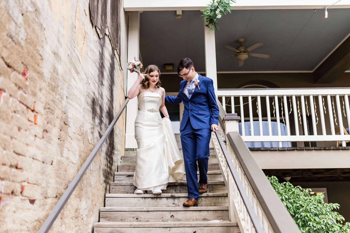A bride in a white dress and a groom in a blue suit walk down wooden outdoor stairs together, holding hands and smiling, with a building and greenery in the background.