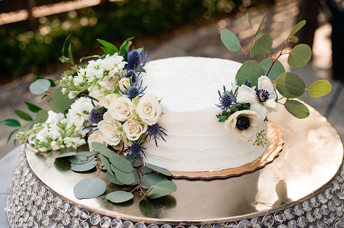Sitting on a crystal cake stand, is a one tier white wedding cake surrounded by white tea roses, silver dollar eucalyptus and blue thistle with accents of anemone.