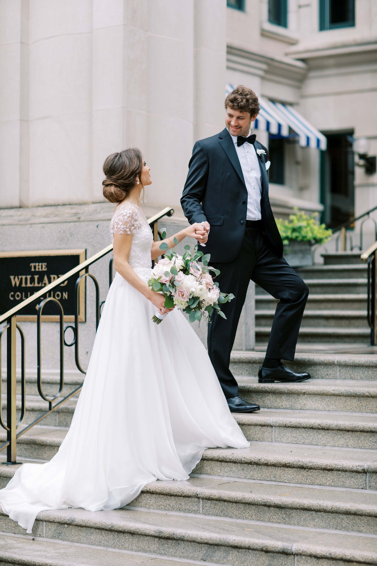 A handsome dark haired groom in a black tux escorts a bride in a white wedding gown holding a bouquet of pink peonies up the stairs of The Willard Hotel in Washington DC as they smile at each other