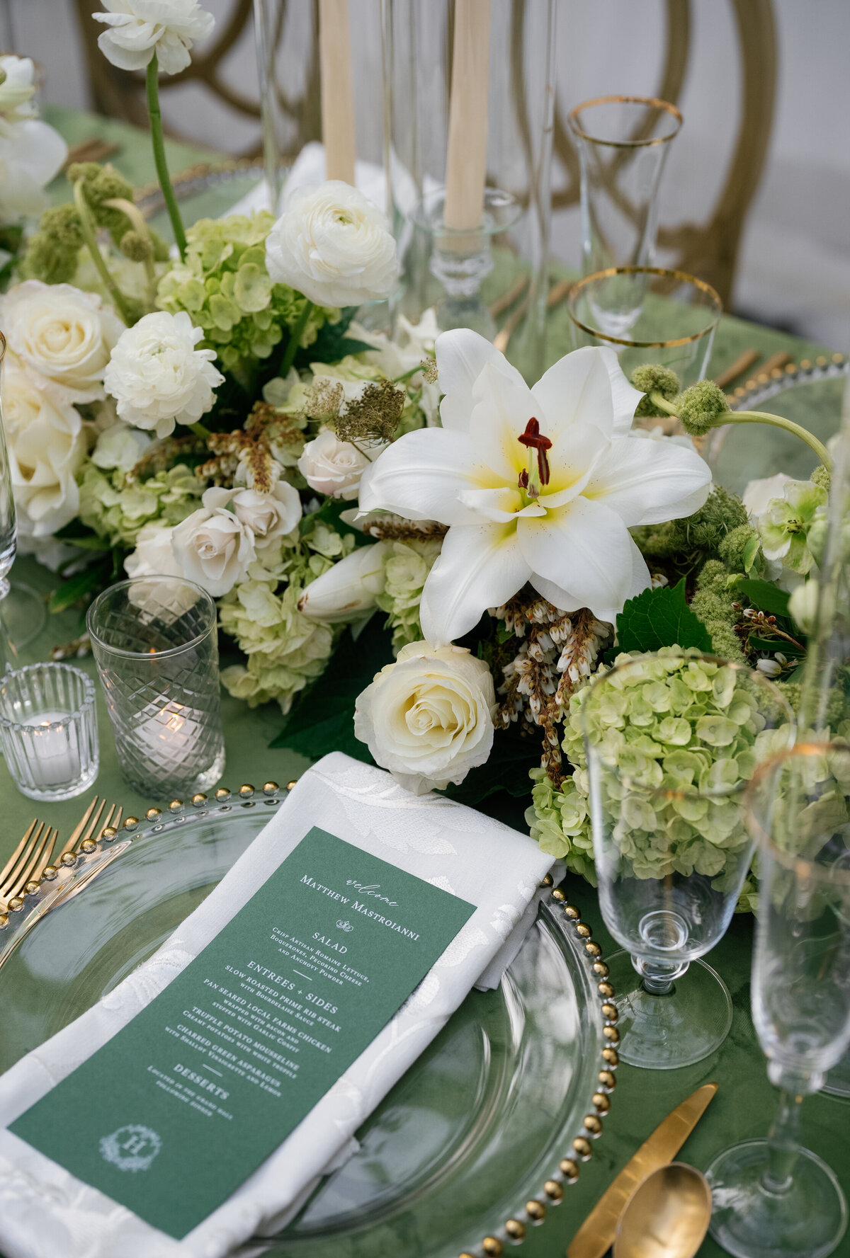 Close-up of a green wedding tablescape featuring a white lily, roses, hydrangea, and gold-rimmed glassware.

