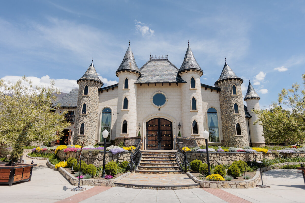 The Castle at Wadley Farms in Lindon, Utah: a cream stone building with six pointed turret towers, arched windows, a round window above carved wooden double doors, and a stone staircase flanked by wrought iron railings and flower beds
