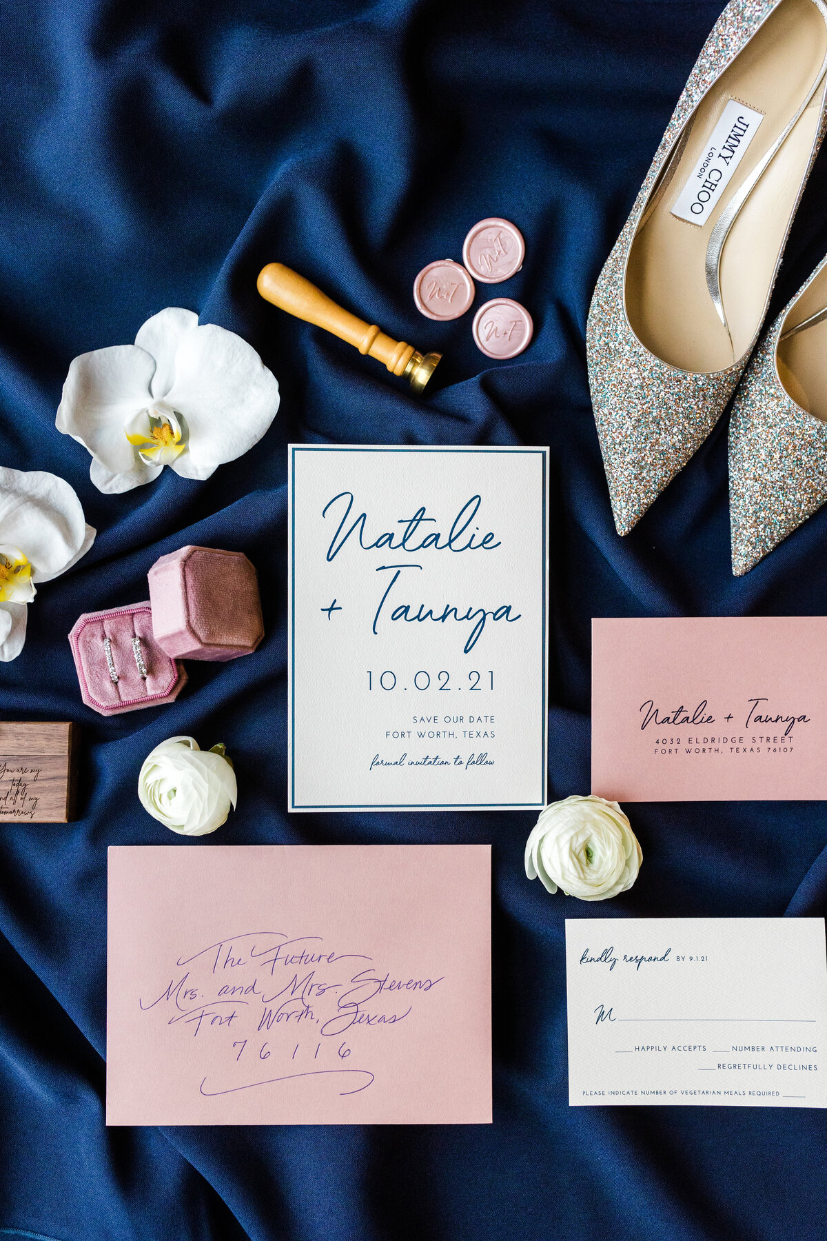 A flat lay detail shot of two brides' invitation, shoes, wax seal and sealer, flowers, rings and their ring box, and RSVP all resting on a dark blue sheet.