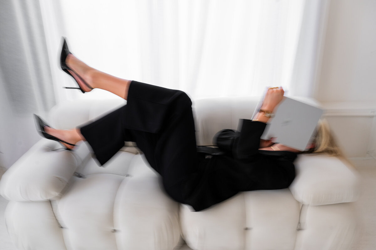 Amelia Waters playfully lounging on a white couch with a laptop covering her face, capturing the fun and relatable side of running a social media agency