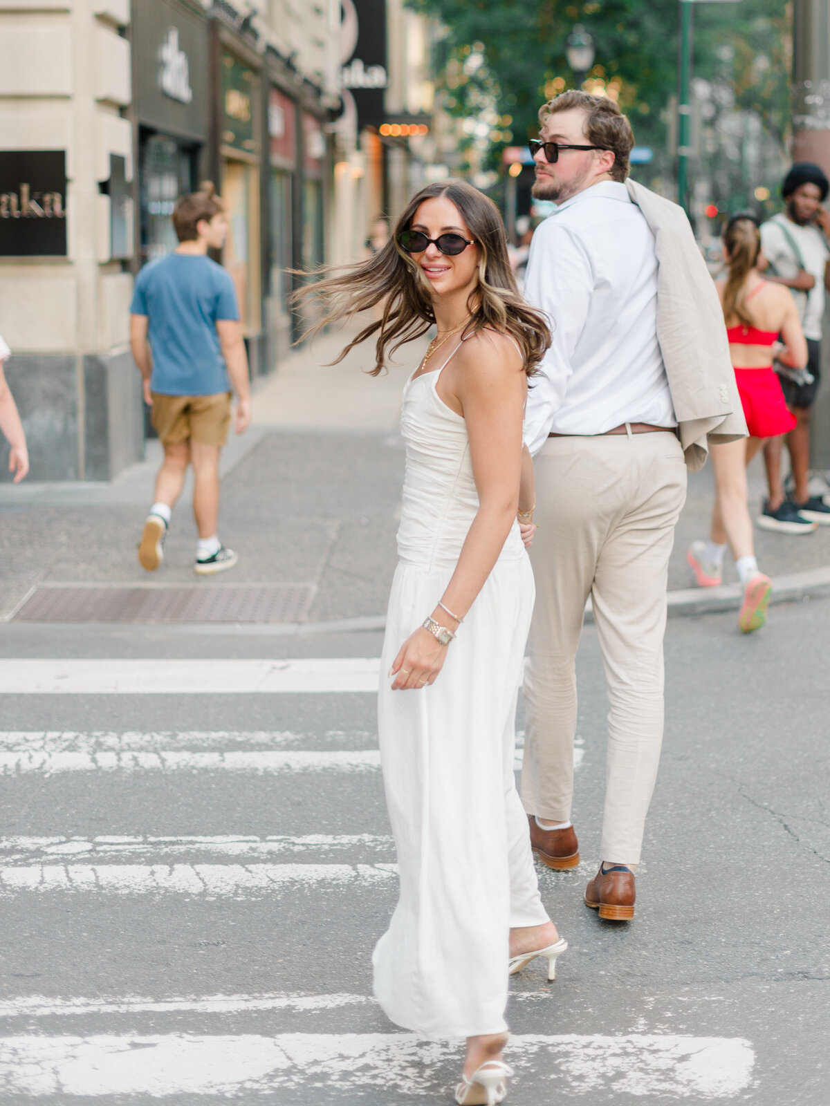 A stylish couple crosses a busy Philadelphia street, exuding confidence and charm. The woman turns back with a radiant smile, her white dress flowing as she walks, while her partner follows with his jacket slung over his shoulder. Captured candidly by Elizabeth Kane Photography, a Luxury Philadelphia Wedding Photographer.