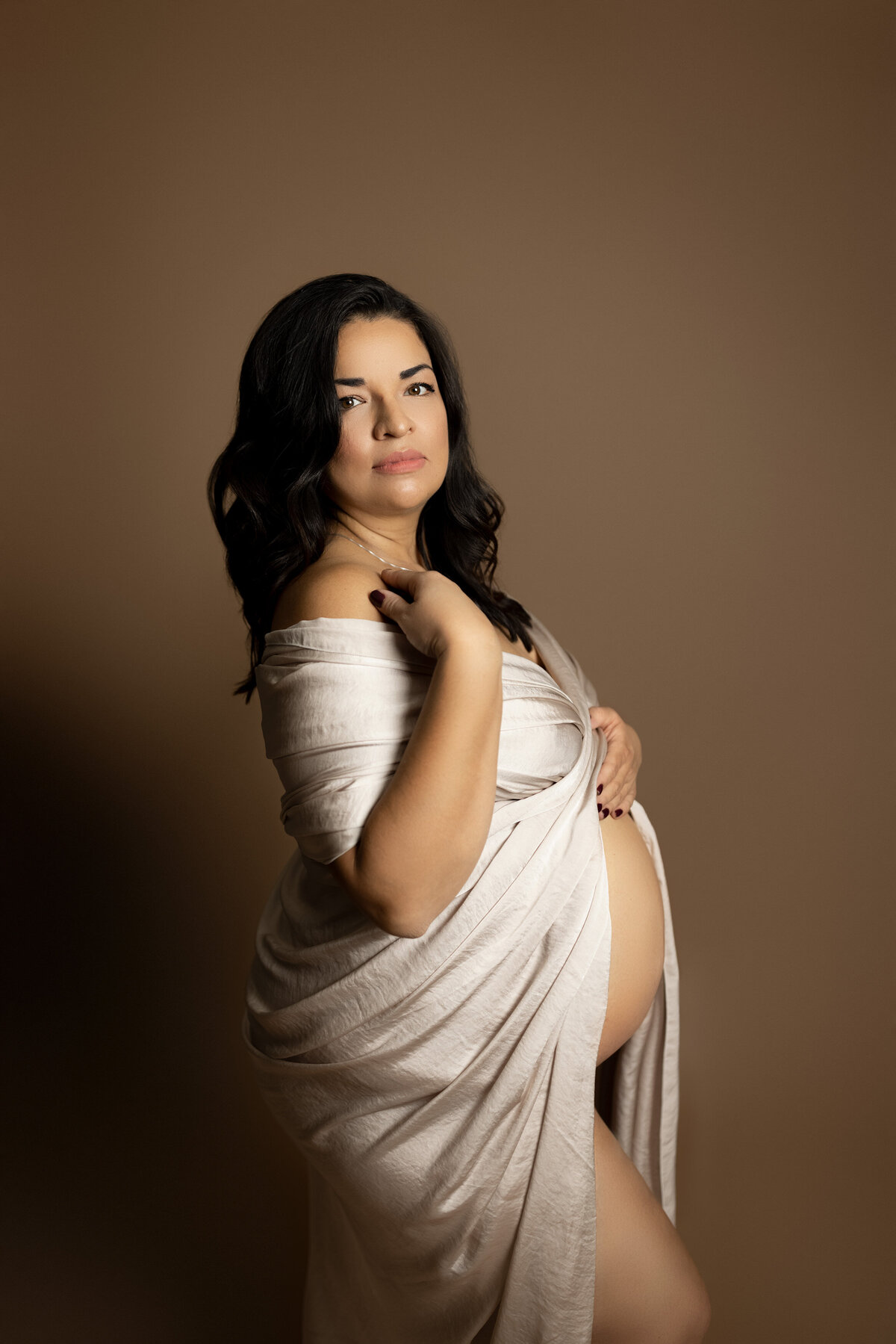 Luxury maternity photoshoot in London, Ontario studio. Mom is draped in a silk gown, but with her belly exposed. Standing side profile to the camera, her front arm is touching her collarbone. She is looking at the camera.