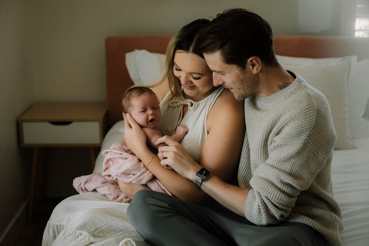 A family of three sitting on the bed while mom holds newborn girl in arms and dad reaches to touch her little hand.