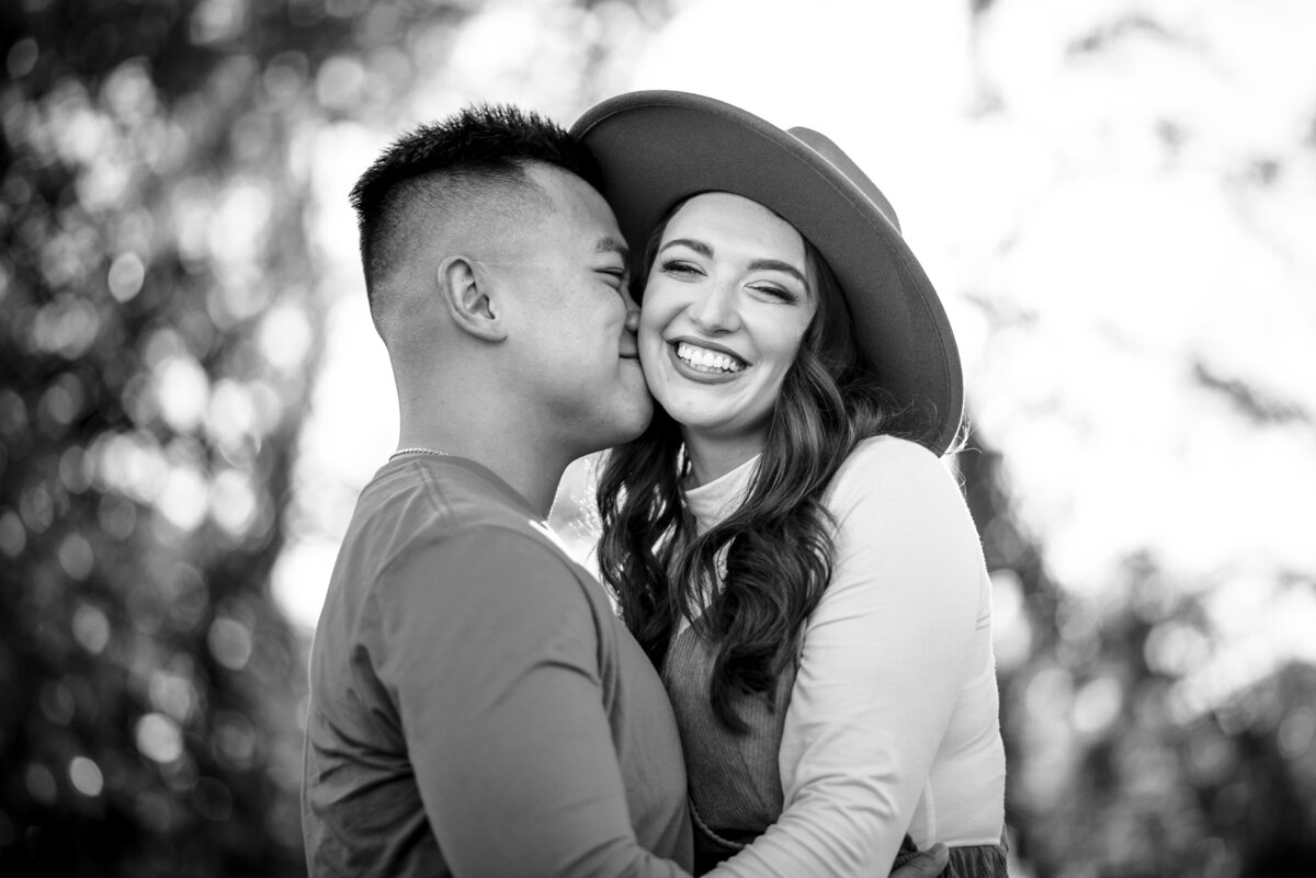 Black-and-white-photograph-of-man-nuzzling-woman’s-temple-while-she’s-giggling-and-looking-at-the-camera-in-a-wide-brimmed-hat-at-Riverfront-Park-in-Columbia-SC