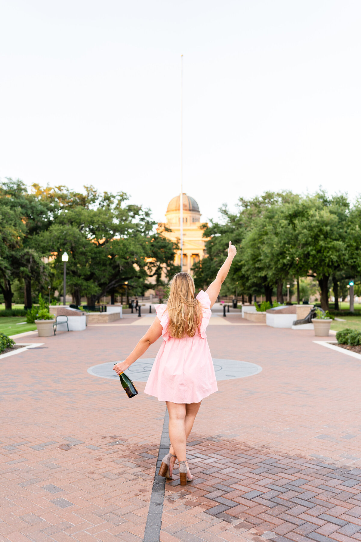 Texas A&M senior girl turned away and holding thumps up while other holds champagne bottle while wearing pink dress at Academic Building in the background