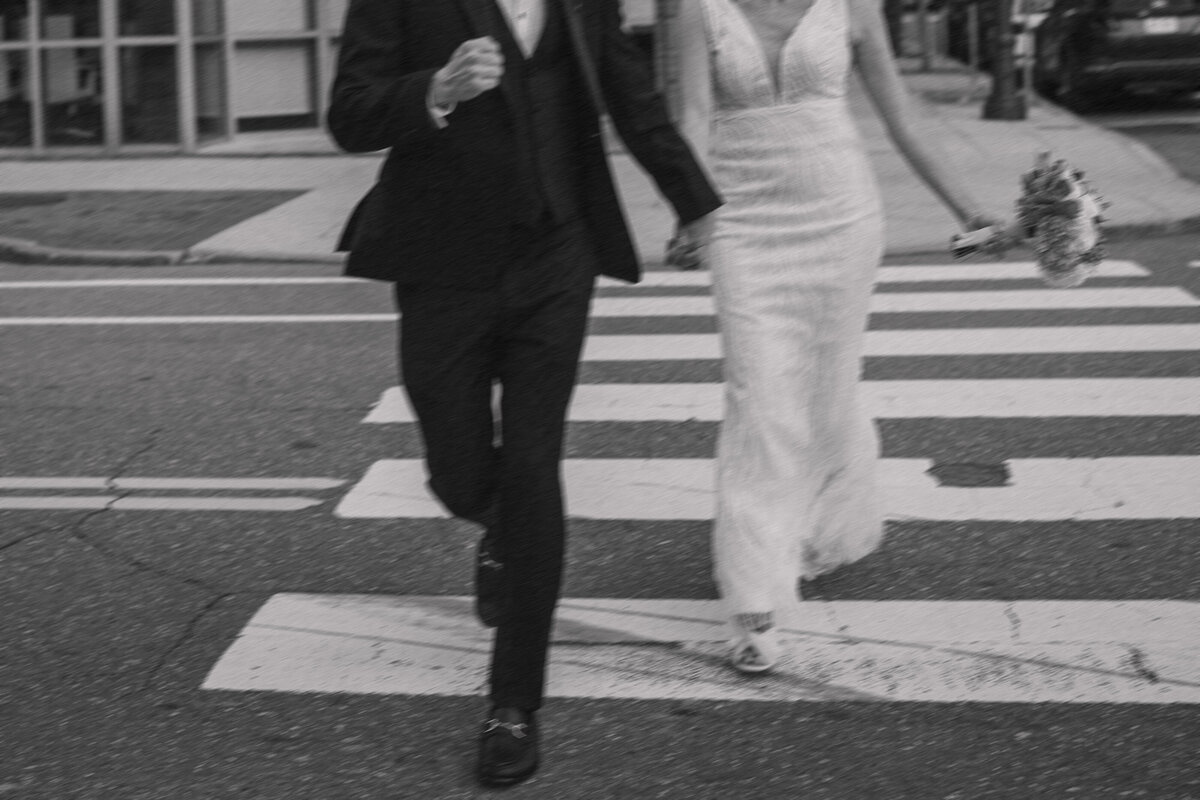 Bride and groom holding hands while running across a city crosswalk. The bride wears a sleek, fitted white dress and holds a bouquet in her free hand, while the groom is dressed in a sharp black suit.