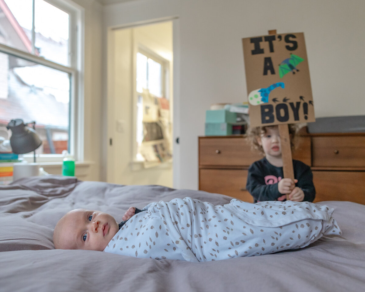 Big sister looks on behind her baby brother laying on the bed