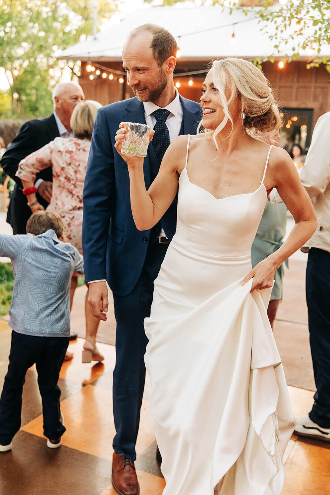 A bride and groom smiling at their wedding reception. The bride is holding a drink in one hand and holds up her dress in the other.