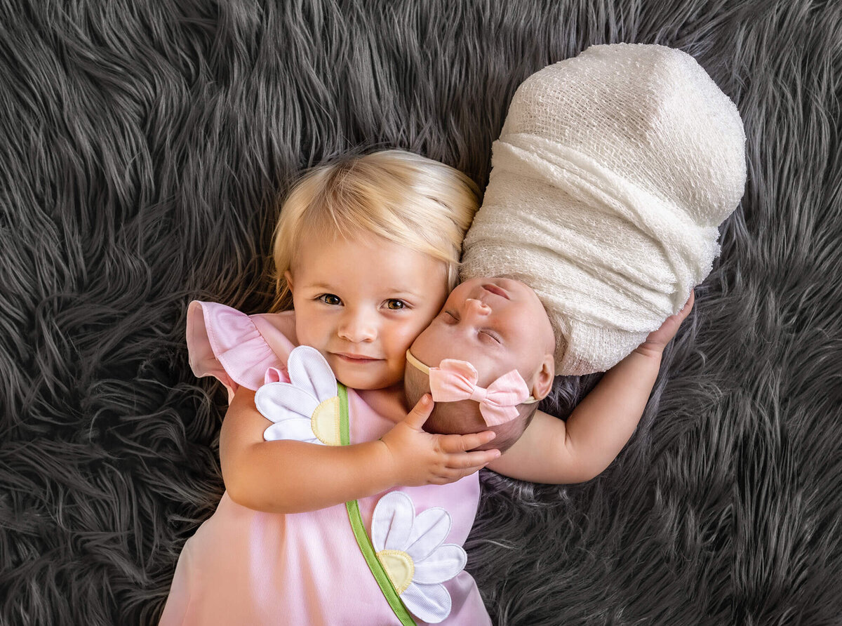 A bib sister cuddles her newborn baby sister who sleeps on her shoulder