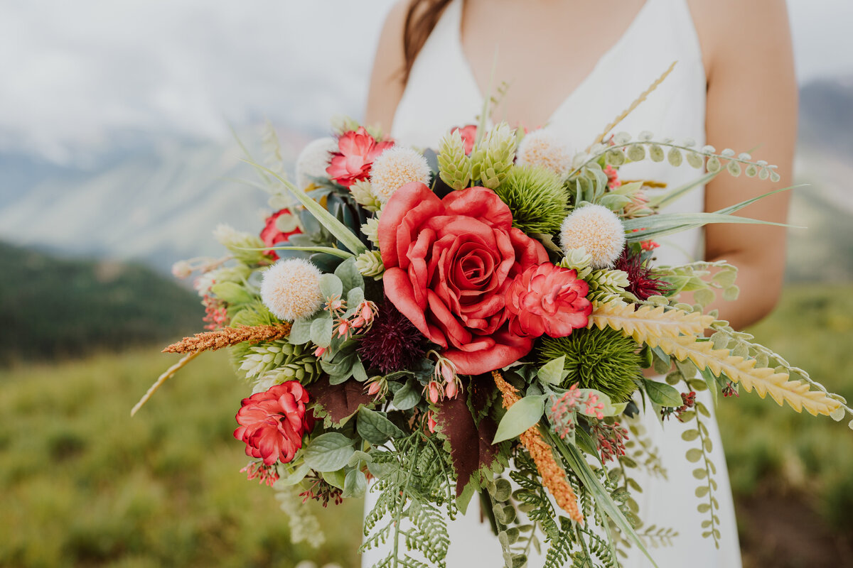 Bride holds a large wedding bouquet with red, white, and green florals on top of a green mountain side in Ouray, Colorado