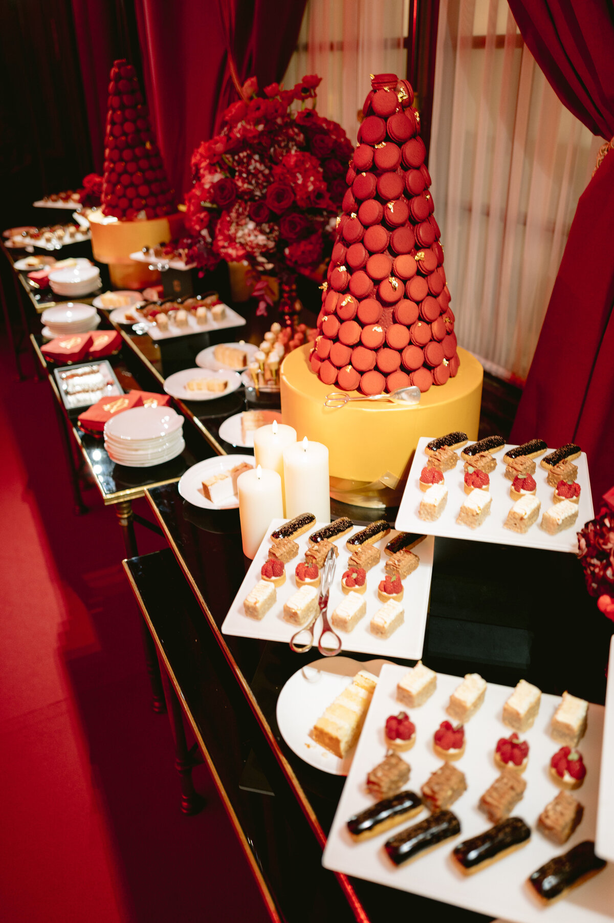 The photo was taken at Shangri La Hotel & Ritz Paris Hotel Paris, France. by photographer Matteo Coltro, with videography by Zen Film Works. It showcases a lavish dessert table featuring a large macaron tower surrounded by various desserts, including mini cakes, chocolate eclairs, and pastries. Candles, red flowers, and ornate decorations enhance the elegant presentation set against rich red drapery.