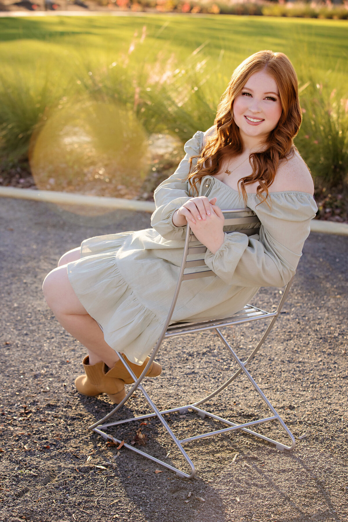 Graceful high school senior in sage green dress sitting in chair with sunflare and layered textures of grass and light