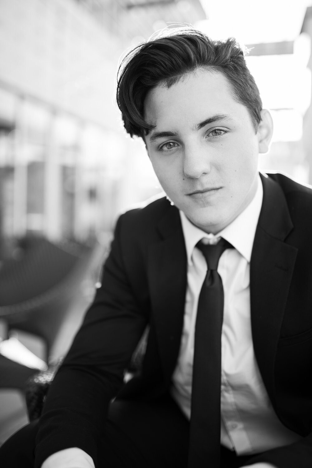 Stylish outdoor senior portrait of a teen boy in a black suit, seated and looking confidently at the camera with editorial flair.