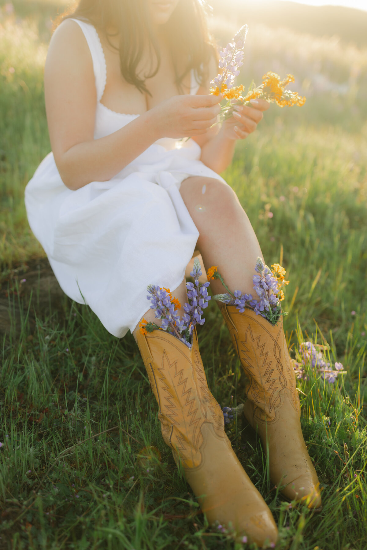 Detail shot of cowgirl boots filled with wildflowers as a woman in a white dress sits in the grass. Captured at golden hour in Greenville, SC by Dani Lacey