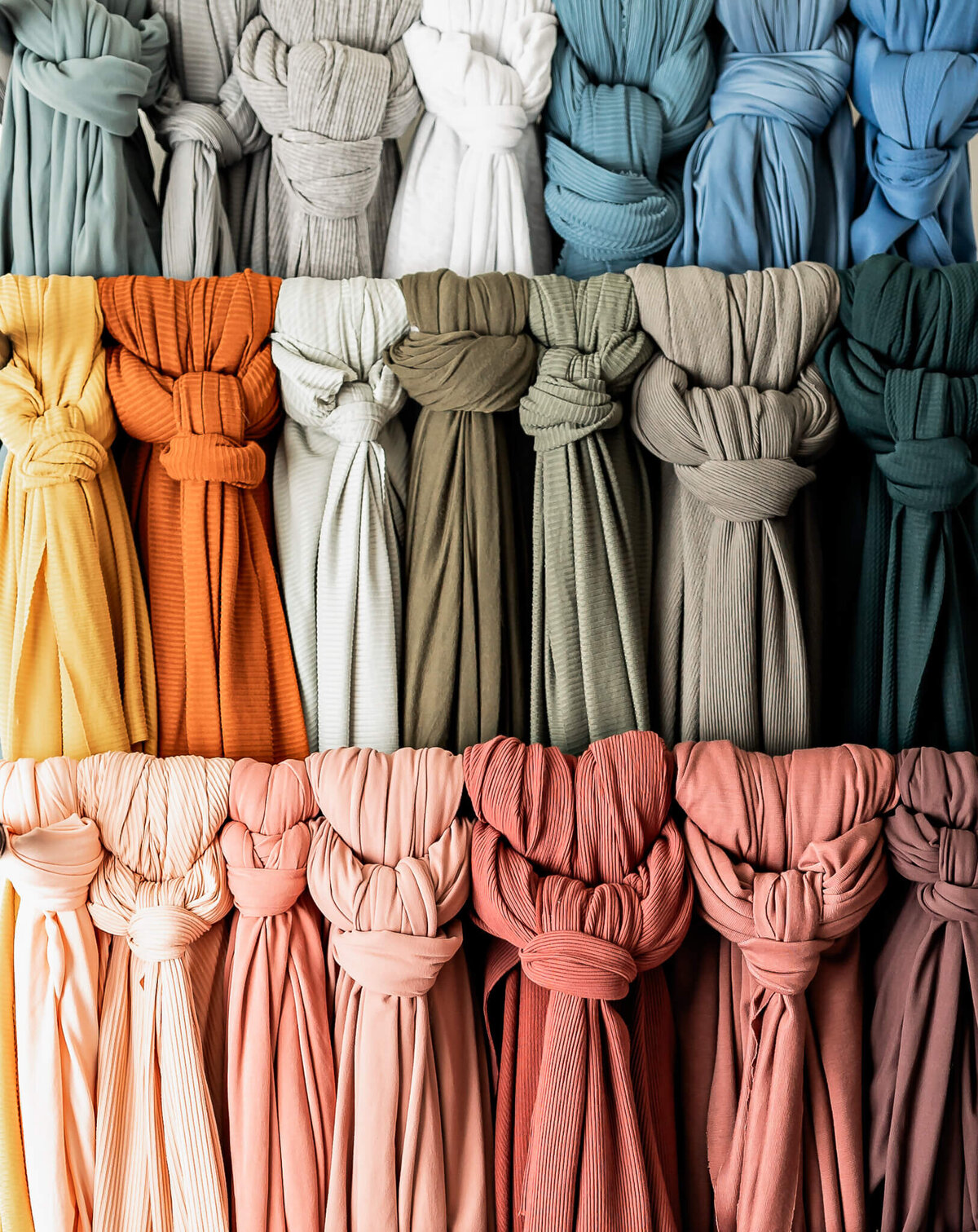 A colorful display of fabric rolls, organized in rows. The top row features blue and white shades, the middle row includes green and neutrals, and the bottom row has orange, pink, and peach tones. Each roll is tied with a large knot.