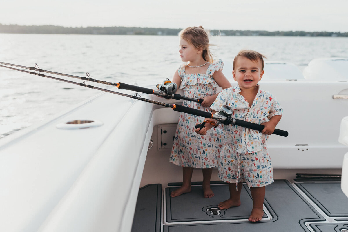 A young brother and sister stand on the back of their family boat holding fishing rods together during their photoshoot with Allison Wolf, Boston Family Photographer