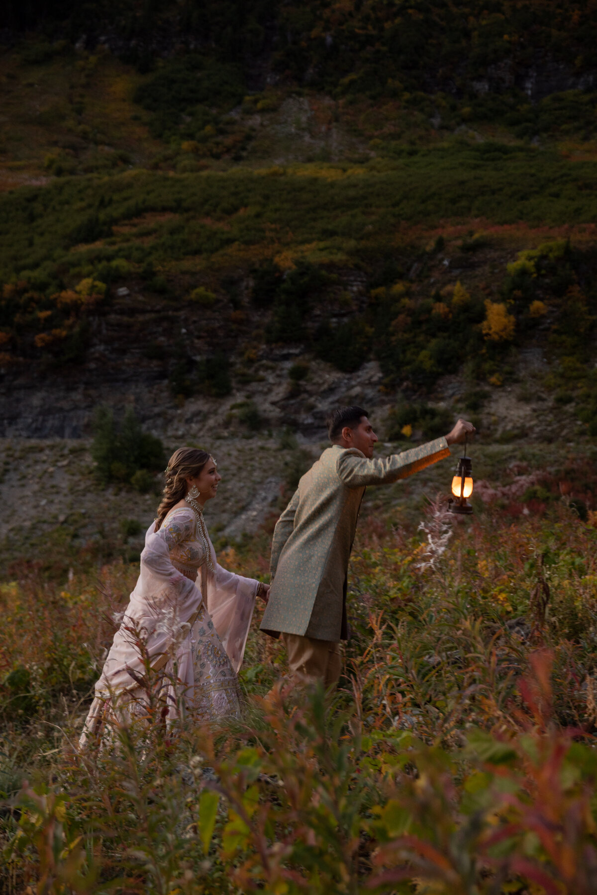 A groom leads his bride through a meadow with a lantern held in front of his face to light the way.