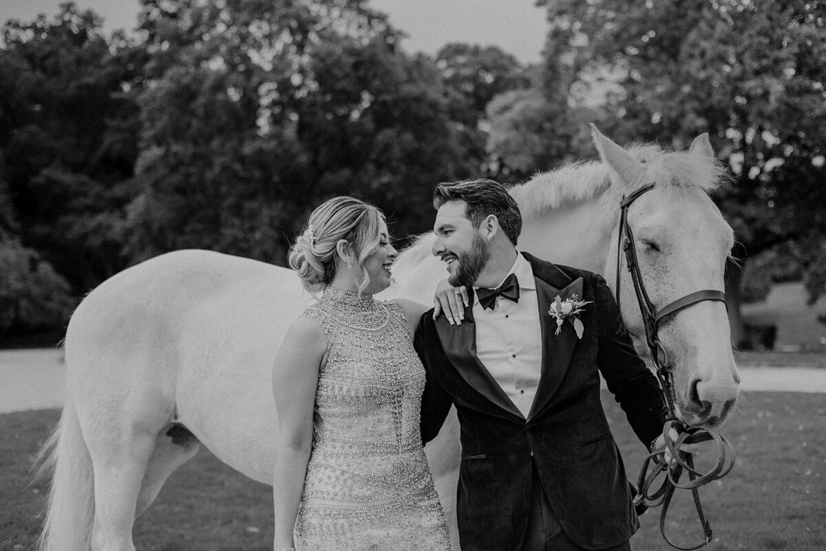 Black and white image of Bride looking at groom holding the reigns of a white horse