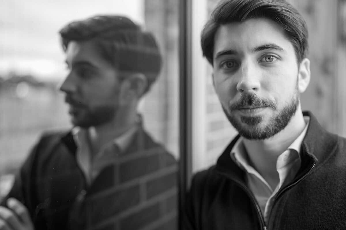 Black and white image of a bearded man leaning against an open door with a reflection of his face in the glass