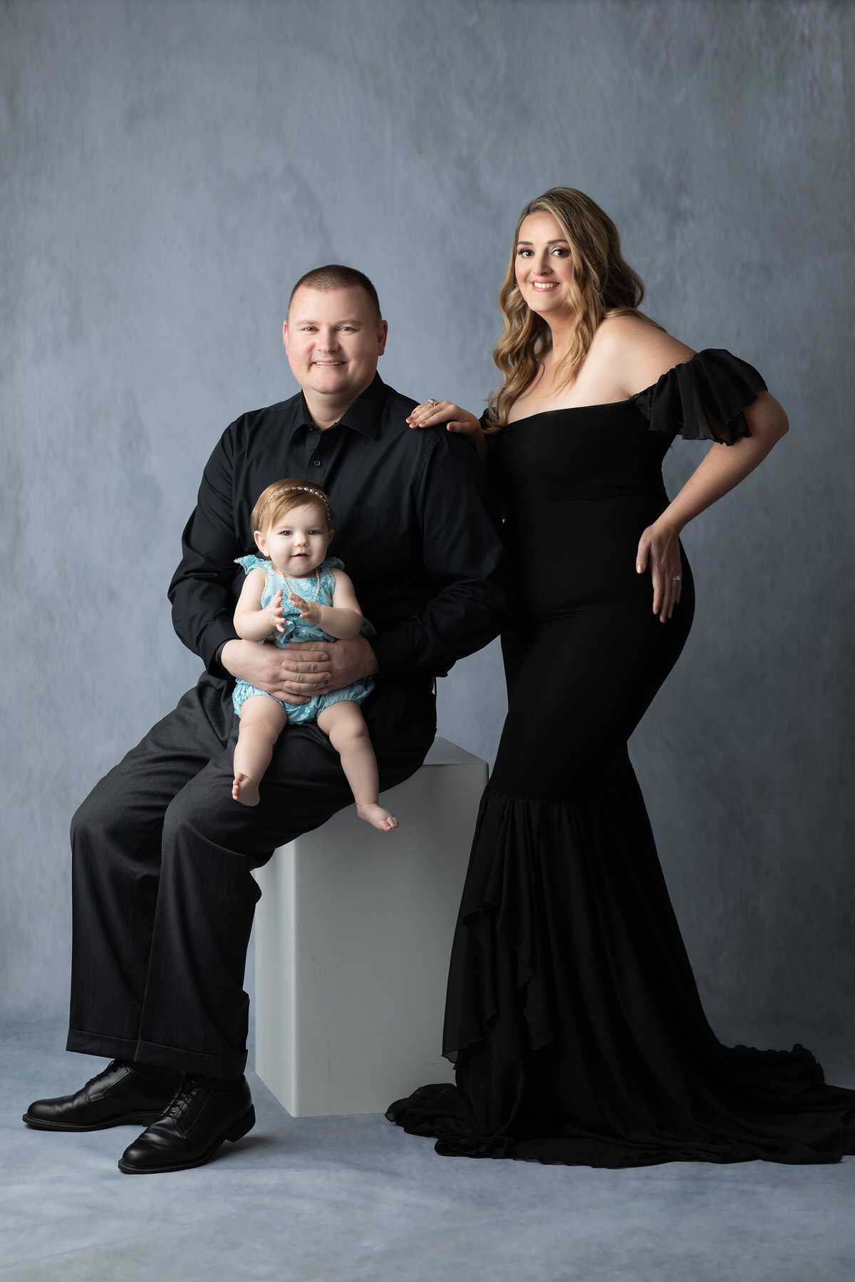 Dallas milestone photographer captures a formal family portrait featuring a mom in a glamorous black gown, dad in classic black, and their baby girl in a teal romper with a sweet smile.