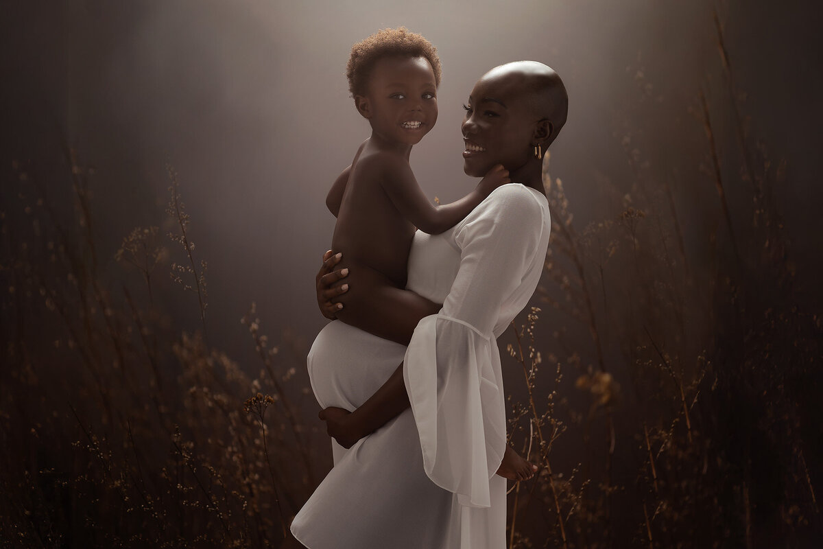 Strikingly beautiful dark skinned expecting mother wearing a short white gown in a flowing fabric holding her toddler son who is sitting on top of her belly.
