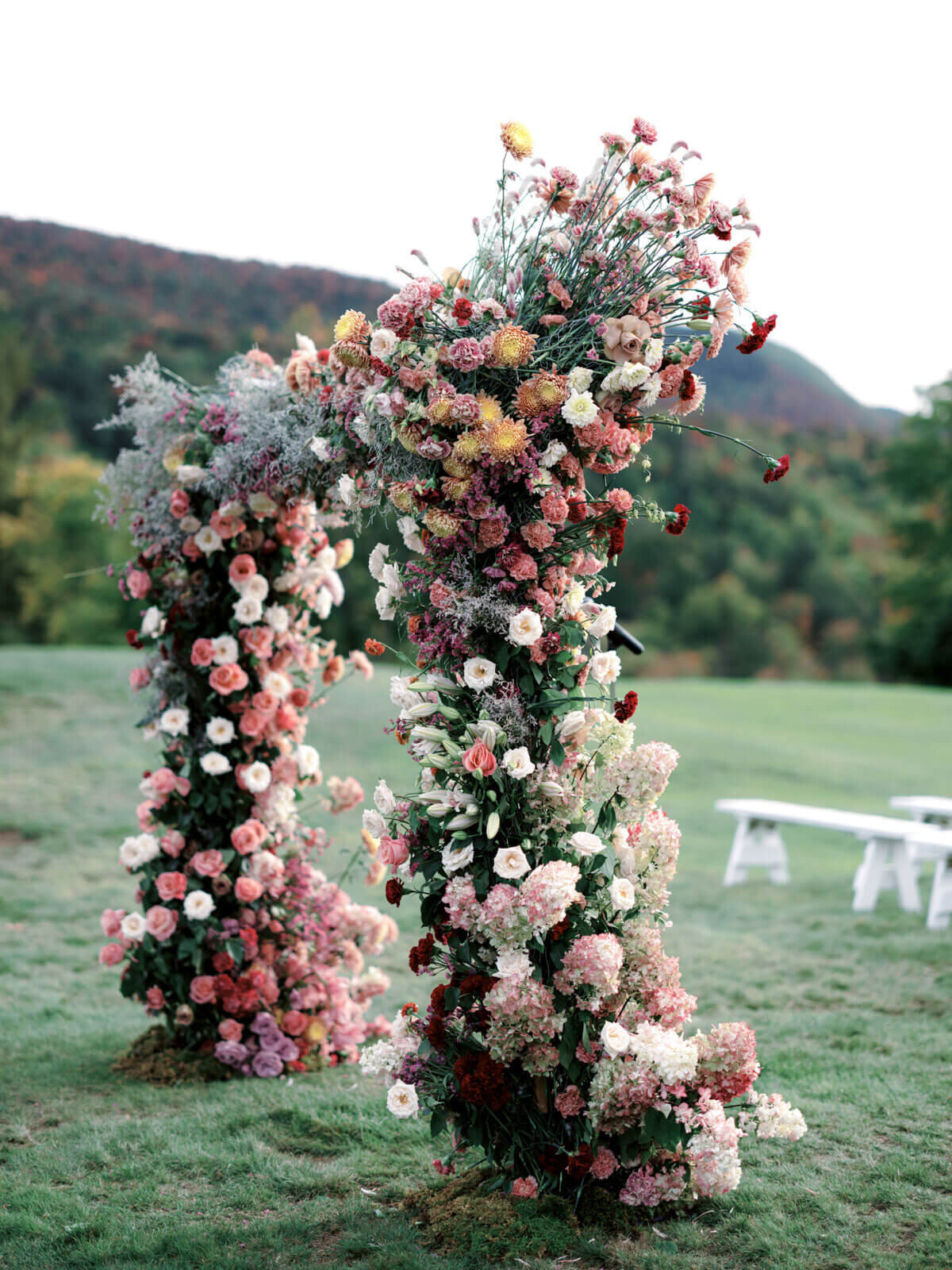 A wedding arch full of peach, pink, red, and white flowers on a golf course at The Ausable Club, NY. Image by Jenny Fu Studio.