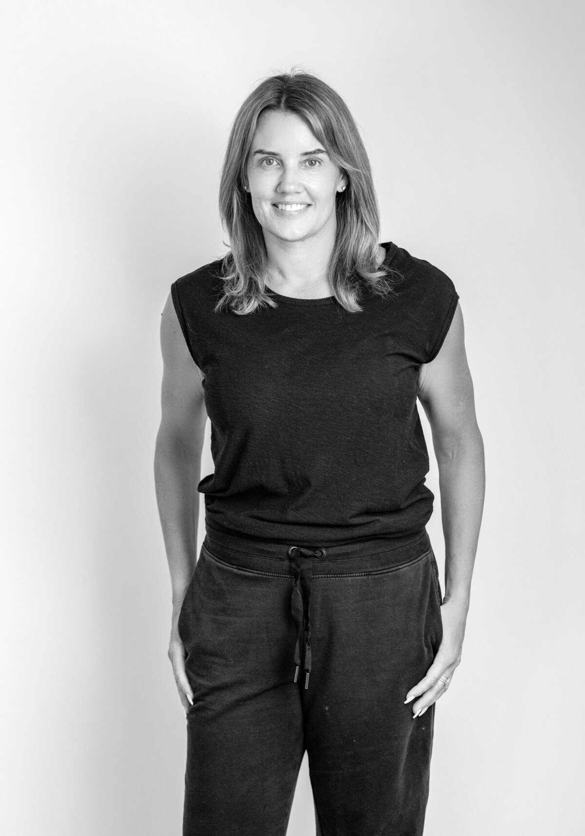 Black-and-white before shot of a woman in a tank top and shorts, smiling confidently with hands on hips.