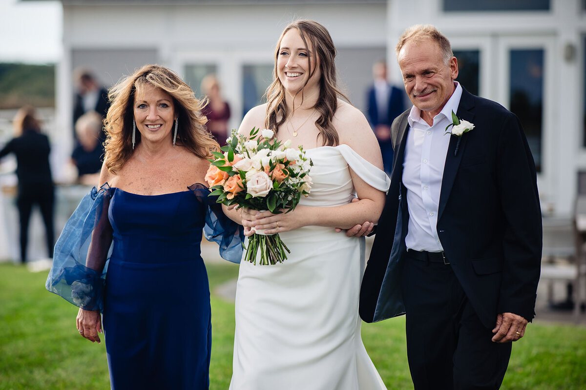 A bride in an off-the-shoulder white dress, holding a bouquet of orange and white flowers, flanked by her parents who are proudly walking with her; her father in a dark suit and her mother in a blue off-the-shoulder dress.
