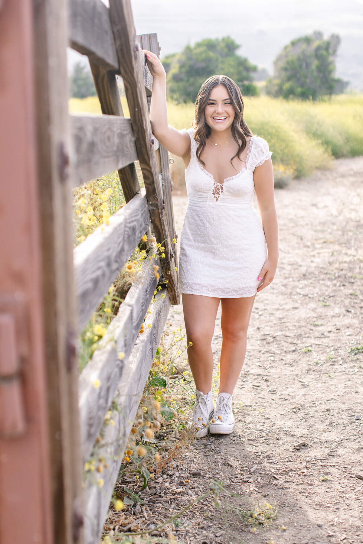 A woman stands by a wooden fence, smiling, in a short white lace dress and white sneakers. The setting is a sunny outdoor path with green grass and trees in the background.