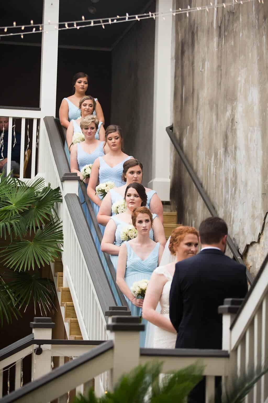 A group of bridesmaids in light blue dresses stand in a line on a staircase behind a bride and groom during an outdoor wedding ceremony in Lincoln Parish, holding white bouquets. String lights hang overhead, and plants are nearby.